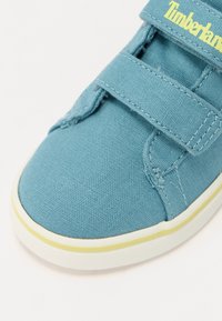 Modrá plátěná bota se zapínáním na suchý zip, bílou podrážkou a žlutým detailem podél okraje podrážky. Na pásku je nápis „Timberland.“