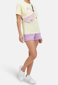 Personne portant un t-shirt FILA jaune pâle, un short lavande, des baskets blanches et un sac bandoulière FILA rose clair sur un fond blanc.