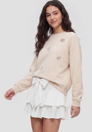 Ung kvinde med langt mørkt hår iført en beige sweatshirt med små dyreansigter og et hvidt lagdelt skørt med en stor sløjfe foran.