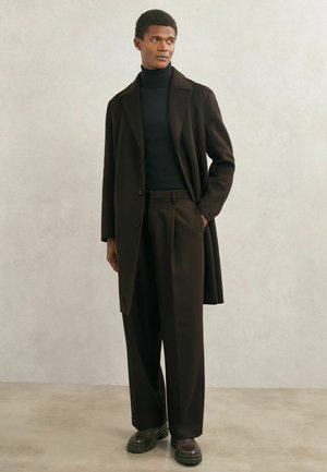 Mann trägt schwarzen Rollkragenpullover, schwarzen Mantel, weite schwarze Hosen und dunkelbraune Stiefel, steht vor einer schlichten hellen Wand.