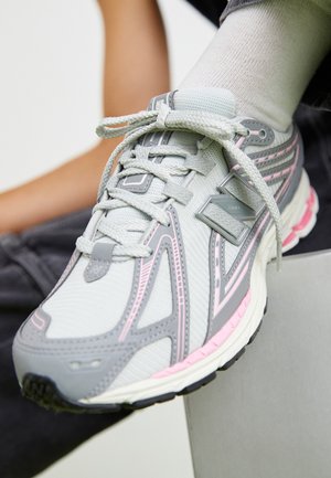 Nærbillede af en grå og lyserød New Balance-sneaker båret med en hvid sok, hvilende på en overflade med en sløret arm i baggrunden.