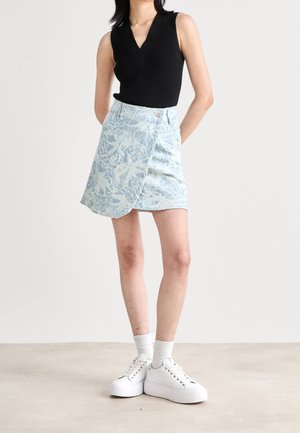 Femme portant un haut noir sans manches à col en V, une jupe portefeuille bleu clair à motifs floraux, des chaussettes blanches et des baskets blanches à plateforme, debout avec les mains derrière le dos.