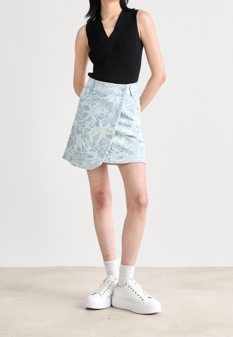 Femme portant un haut noir sans manches à col en V, une jupe portefeuille bleu clair à motifs floraux, des chaussettes blanches et des baskets blanches à plateforme, debout avec les mains derrière le dos.