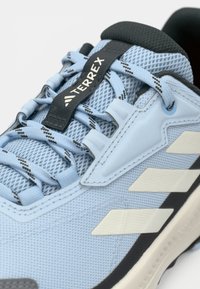 Αθλητικό παπούτσι σε ανοιχτό μπλε από δίχτυ με μαύρες λεπτομέρειες, όπως κορδόνια και σόλα· διαθέτει έντονο λογότυπο και υφασμένη λεπτομέρεια.