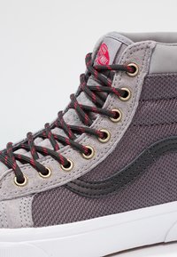 Sneaker typu high-top z szarym zamszem i siateczką, czarnymi gumowymi akcentami, metalowymi oczkami oraz czarnymi sznurówkami z czerwonymi plamkami. Biała gumowa podeszwa.