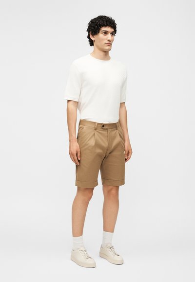 Άνδρας φορώντας ένα απλό λευκό κοντομάνικο πουκάμισο, μπεζ σορτς μέχρι το γόνατο με διπλωμένες άκρες, λευκές κάλτσες και μπεζ αθλητικά παπούτσια, στέκεται μπροστά σε απλό φόντο.