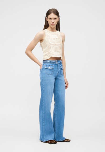 Γυναίκα που φοράει μπεζ αμάνικη πλεκτή μπλούζα, ψηλόμεσο παντελόνι με φαρδύ σκέλος σε μπλε χρώμα και μπροστινές τσέπες, και παπούτσια με μοτίβο λεοπάρ, στέκεται με το ένα χέρι στη μέση.