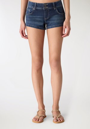 Kobieta w ciemnoniebieskich dżinsowych szortach i beżowych sandałach, stojąca na jednolitym jasnym tle.