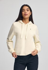 Sudadera color crema con cordón, que presenta un pequeño logo en el pecho, de tela suave y un corte holgado. Se lleva con pantalones oscuros.
