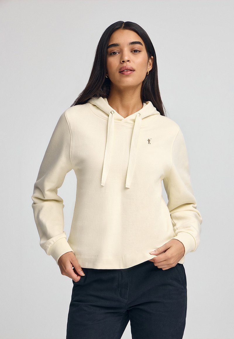 Sudadera color crema con cordón, que presenta un pequeño logo en el pecho, de tela suave y un corte holgado. Se lleva con pantalones oscuros.
