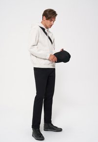 Biała bluza z kapturem, czarny pasek przez ramię, czarne spodnie i czarne sneakersy. Osoba trzyma czapkę w ręku, patrząc w dół, na jednolitym tle.