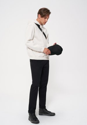 Biała bluza z kapturem, czarny pasek przez ramię, czarne spodnie i czarne sneakersy. Osoba trzyma czapkę w ręku, patrząc w dół, na jednolitym tle.