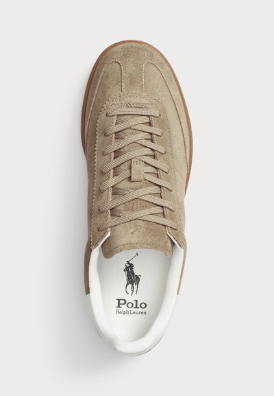 Zapatilla de ante beige con cordones planos, punta redonda y suela de goma marrón en contraste. Presenta un logo impreso en la plantilla.
