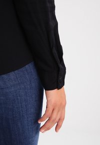 Schwarzes Langarmhemd mit geknöpften Manschetten, kombiniert mit blauer Jeans. Der Stoff wirkt glatt und sitzt an den Handgelenken eng.