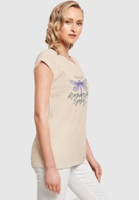 Beige bomulls t-skjorte med korte ermer, med en illustrasjon av en lilla libelle og tekstdesign på forsiden. Vist med hvite bukser.