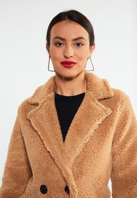 Tan struktureret faux fur-frakke med klippede reverser, dobbeltknaplukning og store sorte knapper. Sort top nedenunder.