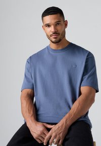 Blå bomulds t-shirt med korte ærmer og rund halsudskæring, prydet med et lille logo på venstre bryst, båret af en siddende person.