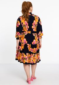 Floral Kleid in Marineblau mit großen rosa und orangefarbenen Mustern. Es hat einen gestuften Rock, lange Ärmel und einen Gürtel in der Taille. Rosa Blockabsätze.