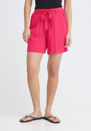 Person trägt leuchtend pinke, lockere Shorts mit gebundenem Bund, schwarzes Oberteil, silberne Ringe und schwarze Flip-Flops vor weißem Hintergrund.