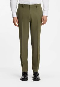 Pantalones hechos a medida color verde oliva, tela suave, parte delantera plana, piernas con pliegues, trabillas para cinturón, combinados con zapatos negros.