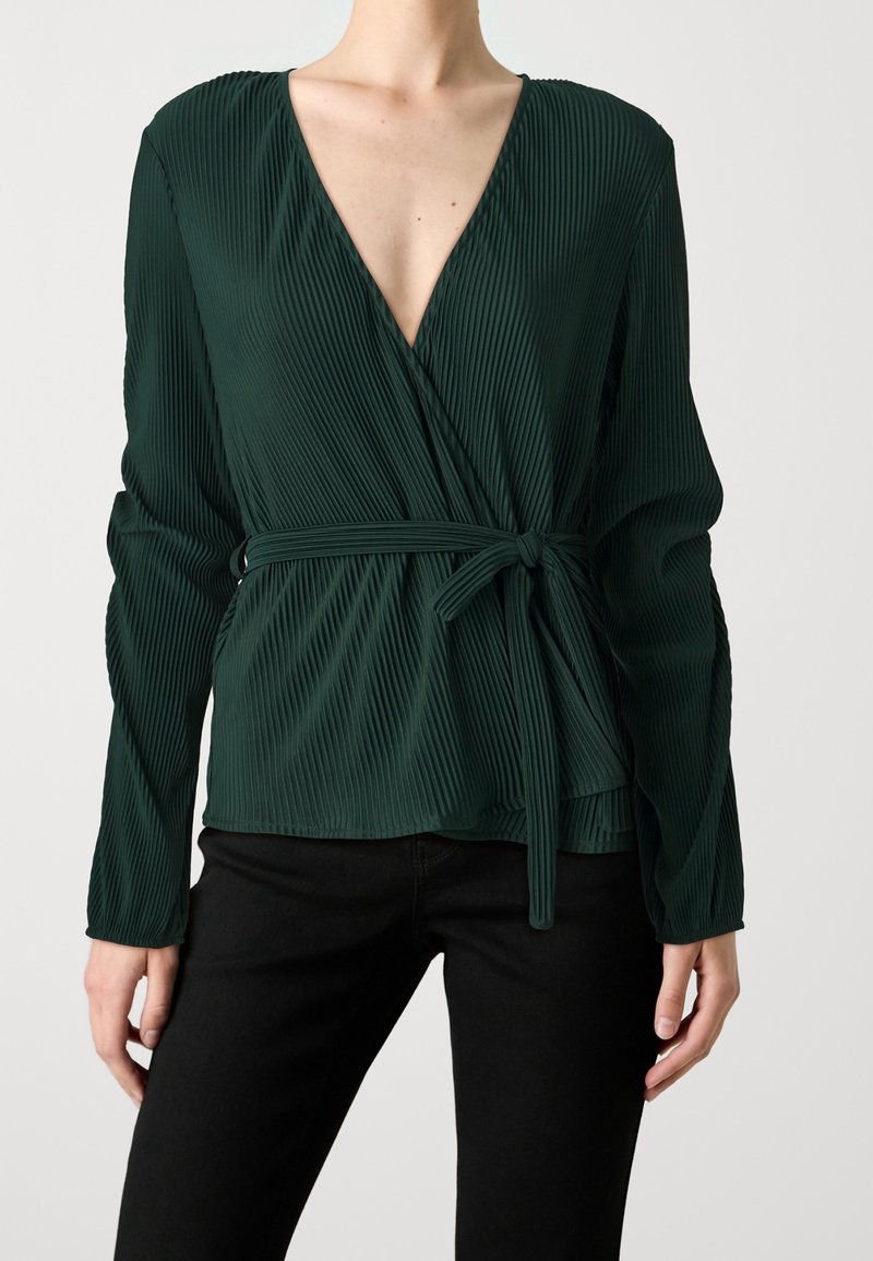 Haut portefeuille plissé vert foncé à manches longues avec un décolleté en V profond et une ceinture nouée à la taille, porté avec un pantalon noir sur une personne debout.