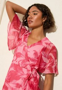 Robe portefeuille rose à motifs floraux avec des manches transparentes et volantes. Dispose d'une taille à nouer et d'un décolleté en V profond. Tissu élégant et fluide avec des motifs audacieux.