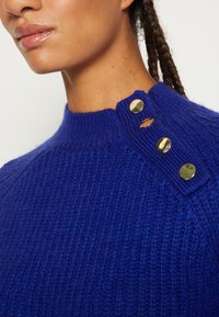 Pull bleu tricoté avec un col montant, orné de trois boutons décoratifs dorés sur l'épaule. Tissu côtelé texturé.