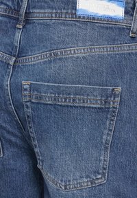 Farkut keskipitkä sininen väri, jossa on hienovarainen pesuvaikutus; siinä on takatasku ja suorakulmainen etiketti vyötäröllä, johon on merkitty bränditiedot.