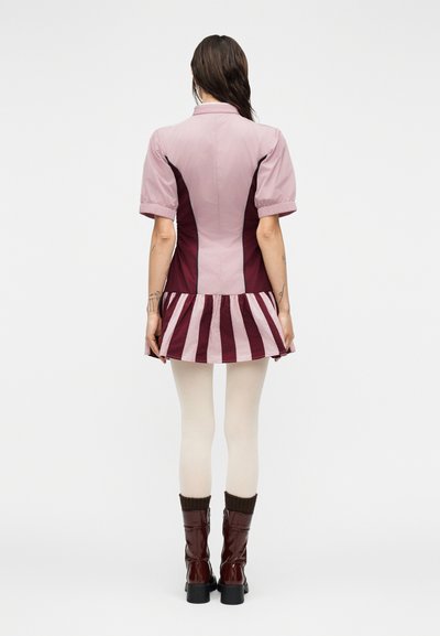 Φόρεμα με κοντά μανίκια σε ανοιχτό ροζ χρώμα με σκούρες μπορντό πλαϊνές πινελιές. Φούστα με ρίγες σε ανοιχτή φαντεζί γραμμή, φορεμένη με κρεμ καλσόν και καφέ γυαλιστερά μποτάκια.