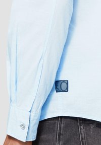  Lichtblauwe blouse met lange mouwen en een knoopmanchet, gemaakt van een textuurstof en voorzien van een zichtbaar zwart etiket met witte tekst.