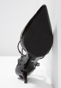 Chaussure à talon haut en cuir verni noir avec bout pointu et brides cloutées à la cheville, vue du côté de la semelle sur fond blanc.