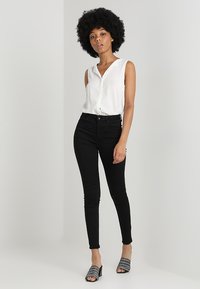 Blouse blanche sans manches avec un devant à boutons, associée à un jean noir ajusté et des sandales à talons ouverts noires avec des accents cloutés.