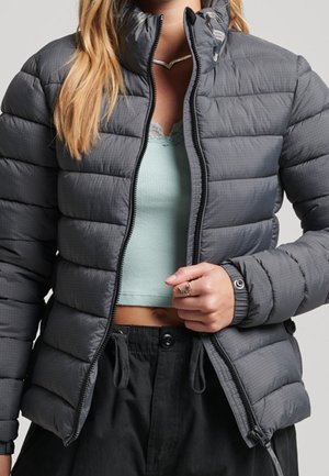 Kobieta w szarej pikowanej kurtce puchowej na lekkim niebieskim krótkim topie i czarnych spodniach, trzymająca suwak kurtki częściowo zapięty.