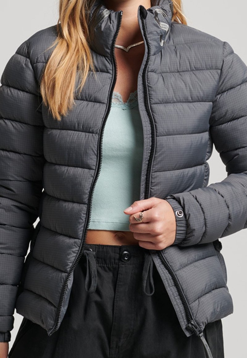 Femme portant une doudoune matelassée grise par-dessus un débardeur court bleu clair et un pantalon noir, tenant la fermeture éclair de la veste partiellement fermée.