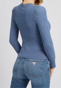 Maglione azzurro lavorato a maglia con un design a tessitura aperta e texture. Orlo e polsini a coste. Abbinato a jeans in denim blu con un logo di marca triangolare.