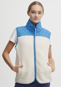 Puffervest med en blå quiltet overdel og en kremfarget fleece underdel. Har en frontglidelås, høy krage og sidelommer.