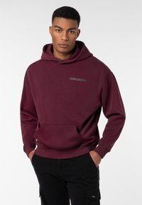 Bordeauxroter Kapuzenpullover mit Fronttasche und dem Text "WORLDWIDE". Weicher Stoff, gerippte Bündchen und Saum, kombiniert mit schwarzen Hosen.