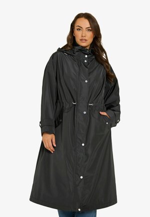 Donna che indossa un lungo impermeabile nero con cappuccio, bottoni a scatto, coulisse in vita e maniche lunghe, in piedi con una mano in tasca.