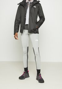 Černá izolovaná bunda na zip, šedé legíny s černými bočními panely a černé turistické boty s růžovými akcenty. Vše na jednoduchém pozadí.