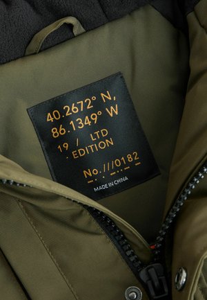Intérieur de veste verte avec une étiquette noire portant des coordonnées et des détails d'édition en texte doré. Tissu lisse avec une fermeture éclair et un bouton-pression.
