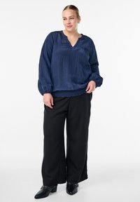 Ženska stoji v temno modri bluzi z dolgimi rokavi, ohlapnih črnih hlačah in črnih čevljih s koničastim nosom ter s sponko, proti prazni beli ozadju.