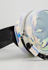 Irydyscentna okrągła torba o błyszczącym wykończeniu, srebrny zamek błyskawiczny, czarny pasek oraz oznaczenie "Steve Madden" na metalowych elementach. Teksturowana powierzchnia z efektem błysku.