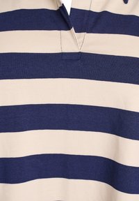 Polo majica s prugicami, ki vključuje široke mornarsko modre in bež vodoravne črte, bombažna tkanina, kratka vratna odprtina in fine teksture.