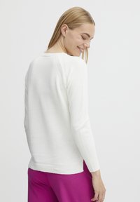 Pull en tricot blanc à manches longues, avec des poignets et un ourlet côtelés, présentant un col rond et des coutures d'épaule discrètes. Porté avec un pantalon rose vif.