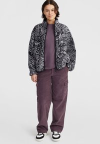 Veste en polaire avec des motifs géométriques noir et blanc, fermeture éclair ; associée à un pantalon cargo en velours côtelé violet foncé et des baskets noir et blanc.