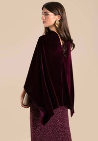 Una chalina de terciopelo burdeos se despliega sobre los hombros, con un diseño suelto y fluido. Combinada con un vestido púrpura estampado y un pequeño bolso de mano plateado.