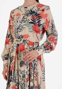 Florales Kleid in Beige mit roten und marineblauen Mustern. Lange Ärmel, runder Ausschnitt und ein Taillengürtel. Leichter Stoff mit gerafftem Rock.
