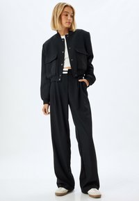 Veste bomber noire raccourcie avec des boutons pression, deux poches à l'avant et des poignets côtelés, associée à un pantalon noir ample et des sneakers blanches.