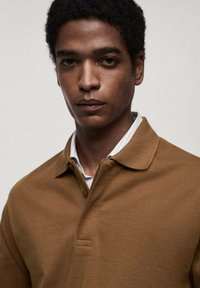 Hnědý polo tričko se strukturovaným límcem a třemi knoflíky, vyrobené z hladkého, texturovaného materiálu; nošené přes košili s bílými pruhy.