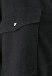 Mustapintainen kangas, jossa on teksturoitu pinta, ja tasku hopeanvärisellä nepparilla sekä ommelluilla yksityiskohdilla.